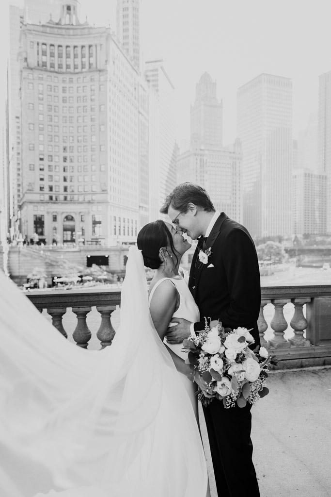 Black and white photo of bride and groom embracing near the Wrigley Building in Chicago with downtown buildings in the background, the bride's veil flowing in the wind