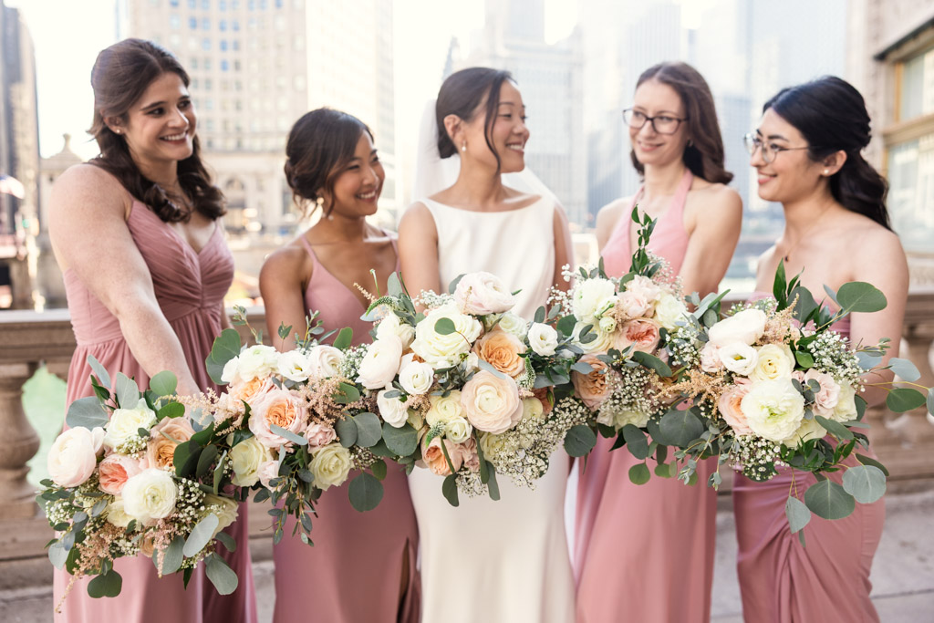 A bride and four bridesmaids in pink dresses smile and hold bouquets outdoors near the Wrigley Building, ready to celebrate at Salvatore's