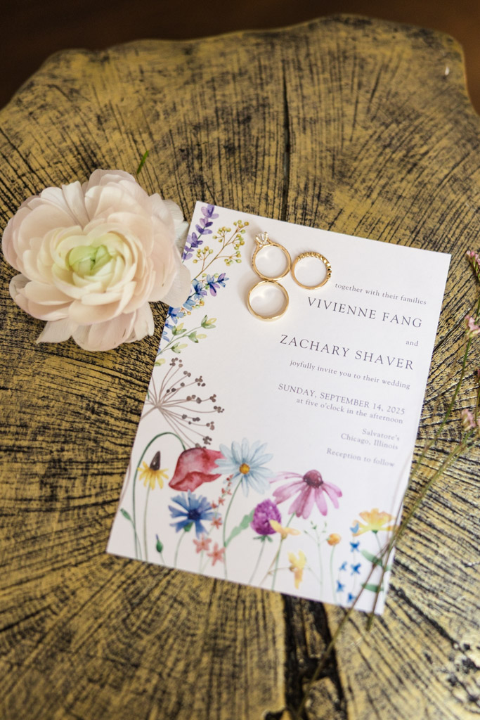 Wedding invitation with floral design, three rings, and a white flower on a textured wooden surface