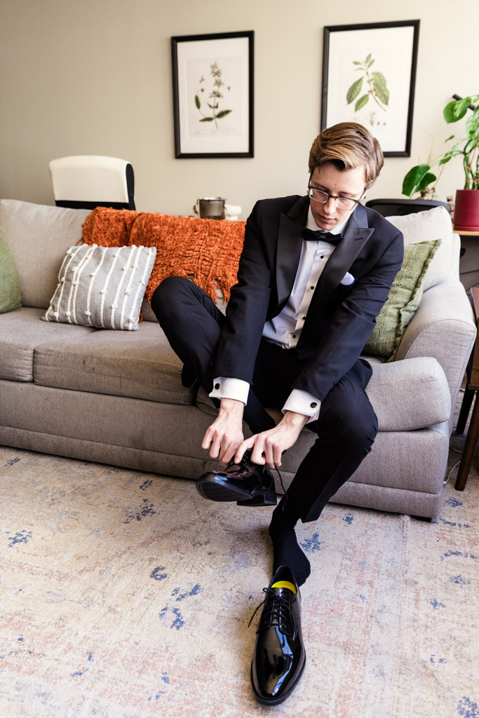 Groom in a tuxedo sits on a couch in the living room, putting on his black dress shoes before his wedding at Salvatore's in Chicago