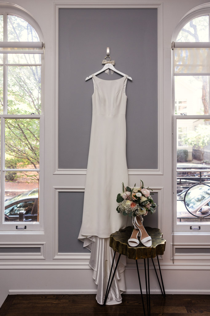 A white sleeveless wedding dress on a hanger, with a bouquet and white heels arranged on a small table below