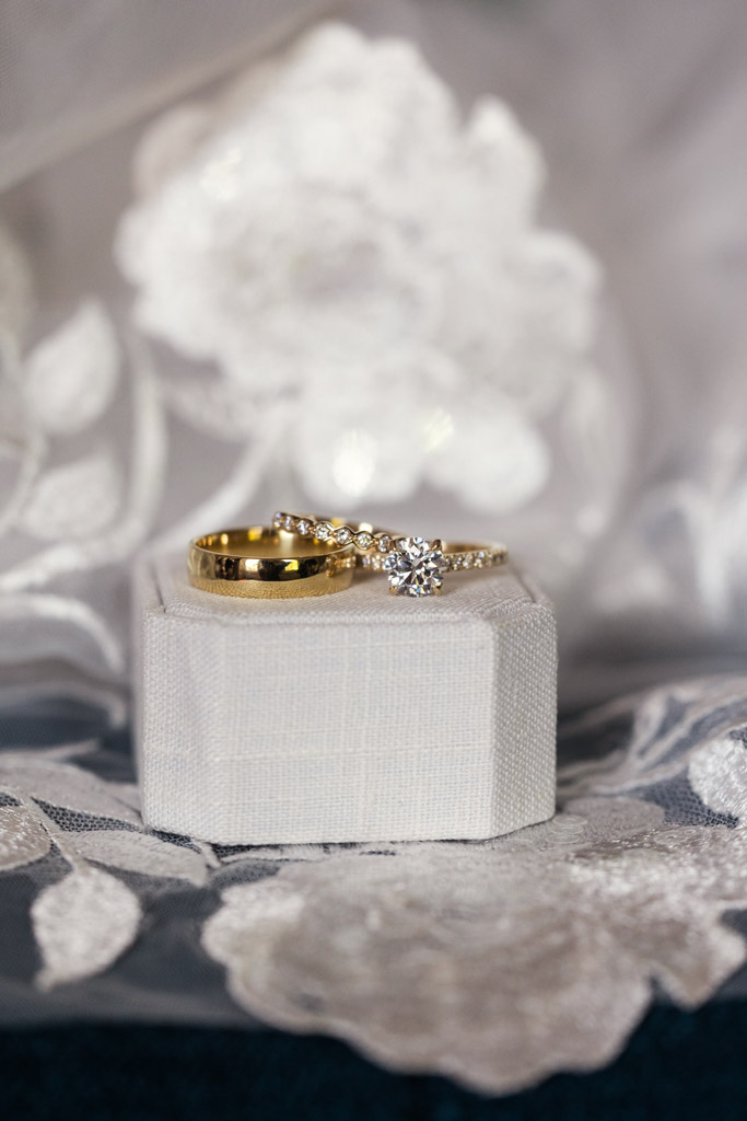 Gold wedding band and diamond ring are elegantly displayed on a white ring box with a floral lace background