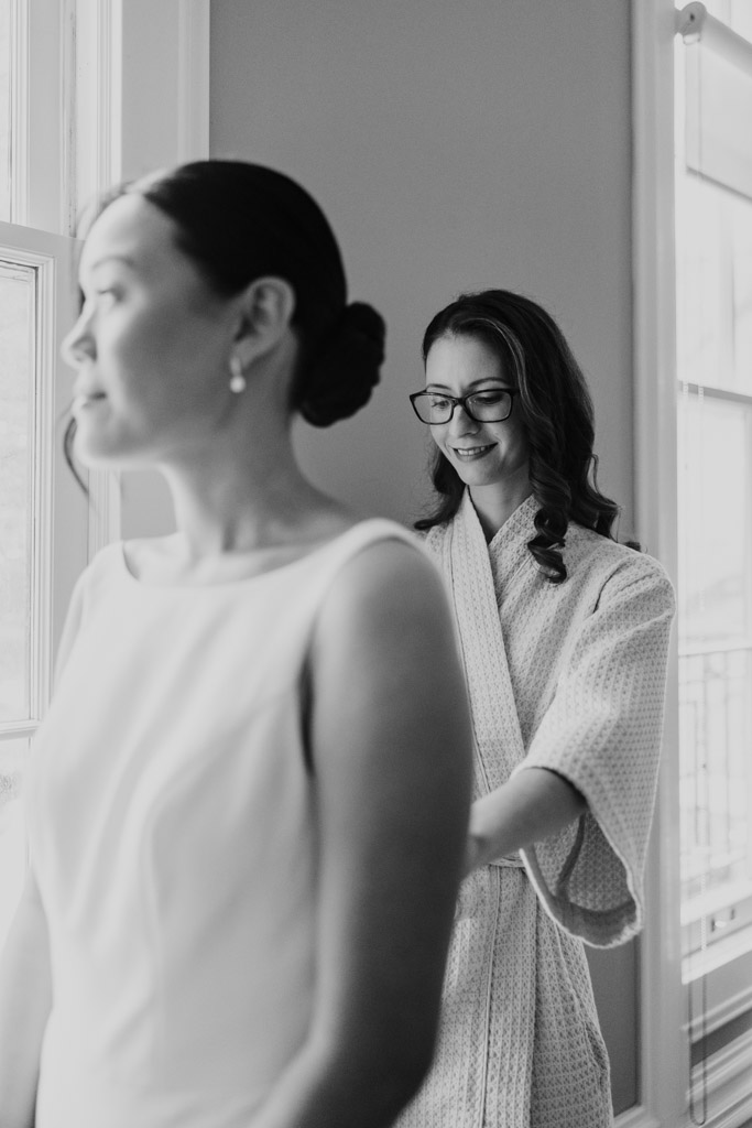 Black and white photo of woman helping the bride adjust her dress near a window, preparing for her wedding at Salvatore's