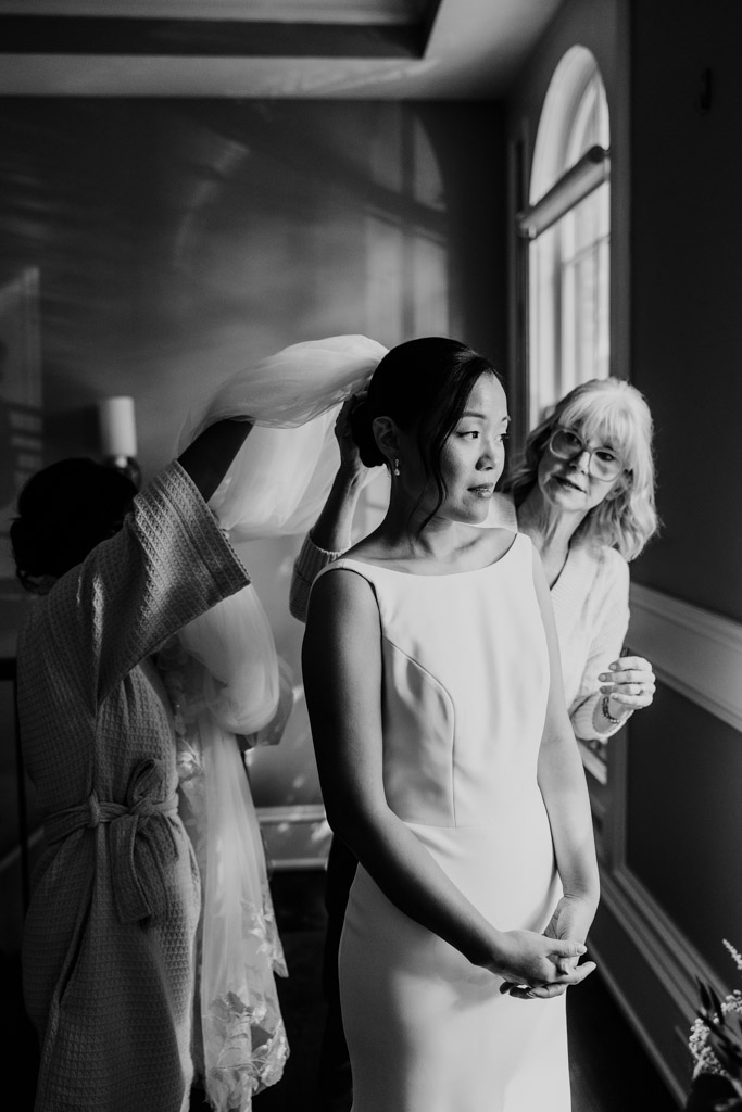 A bride stands in a softly lit room at Salvatore's while two women gently help her with her veil.