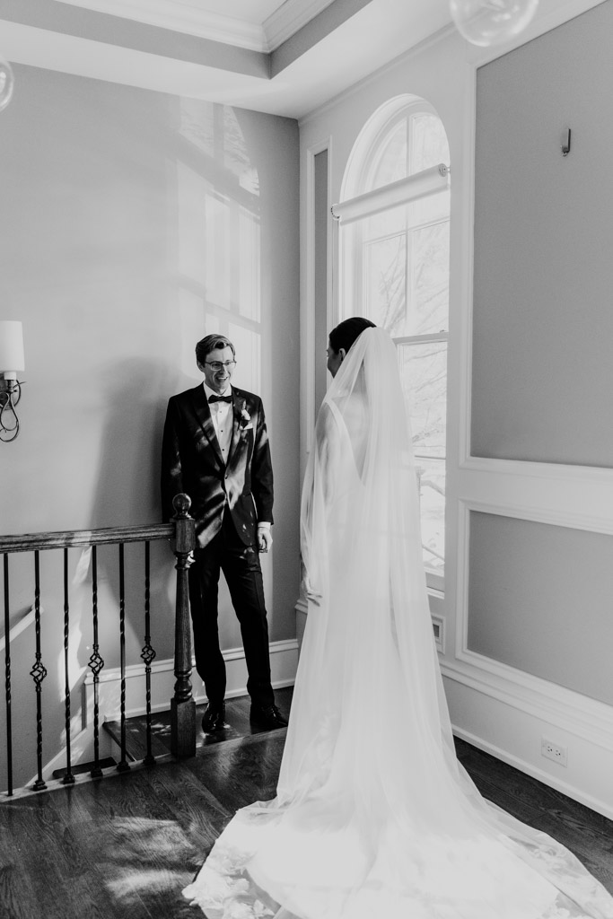 Black and white photo of bride in a long veil \ facing her groom by a window during their first look