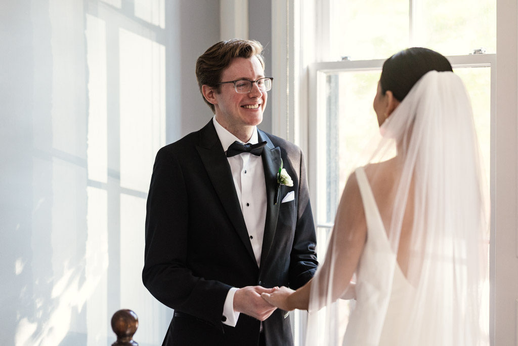 Happy groom in a tuxedo smiles at his bride in a white dress and veil as they hold hands by a window during their first look