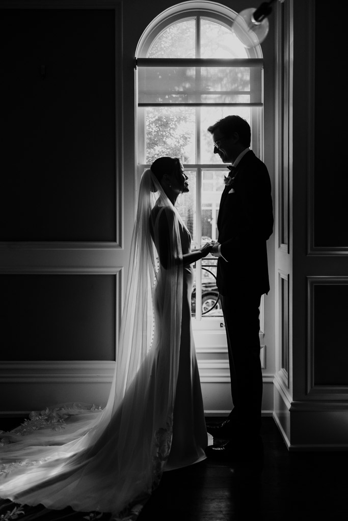 Black and white photo of bride and groom holding hands in front of a window during their first look, silhouetted in soft light