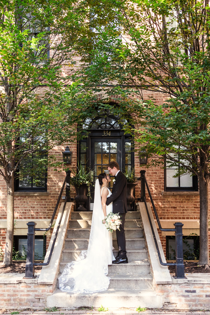 A bride and groom stand together on the outdoor steps of a brick building, surrounded by trees