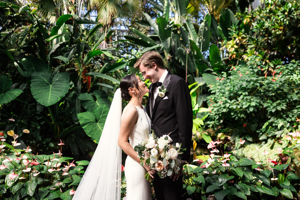 A bride and groom smile at each other in a lush garden at Lincoln Park Conservatory, surrounded by tropical plants and vibrant flowers