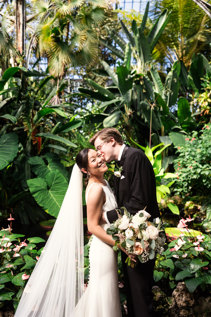 Groom kisses bride's cheek in a lush garden at Lincoln Park Conservatory