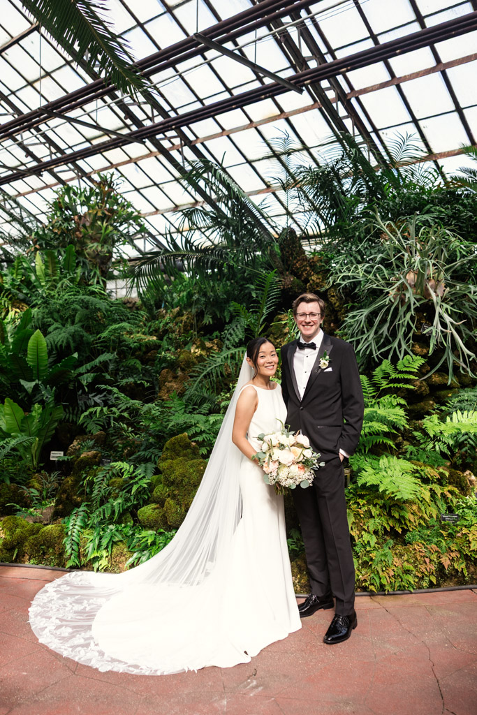 Portrait of ride and groom smiling together in a lush greenhouse at Lincoln Park Conservatory, surrounded by vibrant green plants and ferns