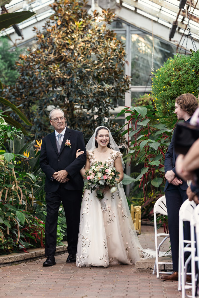 A bride in a white floral gown walks down the aisle with her father in a suit during Lincoln Park Conservatory wedding ceremony