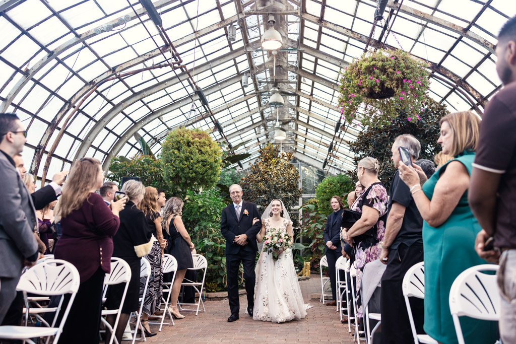 Bride in floral gown walks down the aisle at a Lincoln Park Conservatory wedding, escorted by her father, with guests watching