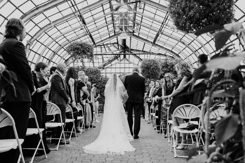 Black and white photo of bride walking down the aisle with her father during her Lincoln Park Conservatory wedding ceremony, guests seated on both sides