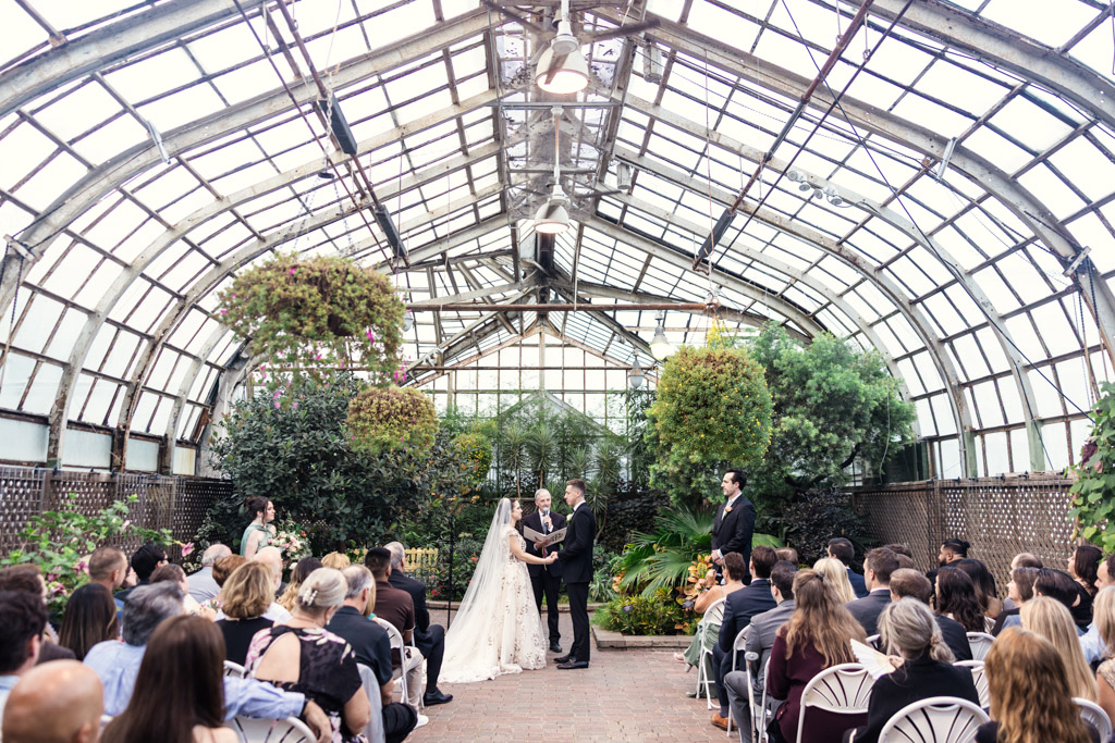 A couple exchanges vows in a greenhouse at their Lincoln Park Conservatory wedding, surrounded by guests seated on either side of the aisle.