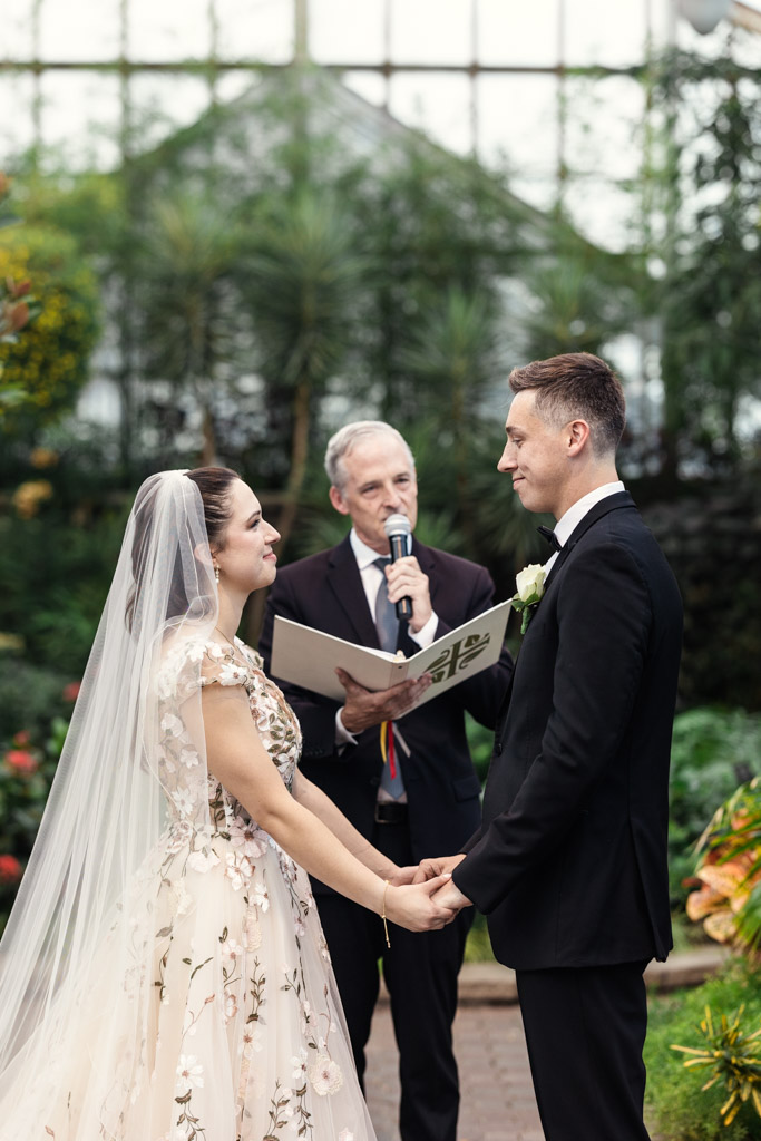 A bride and groom hold hands during a Lincoln Park Conservatory wedding ceremony, with an officiant speaking behind them in a lush garden setting