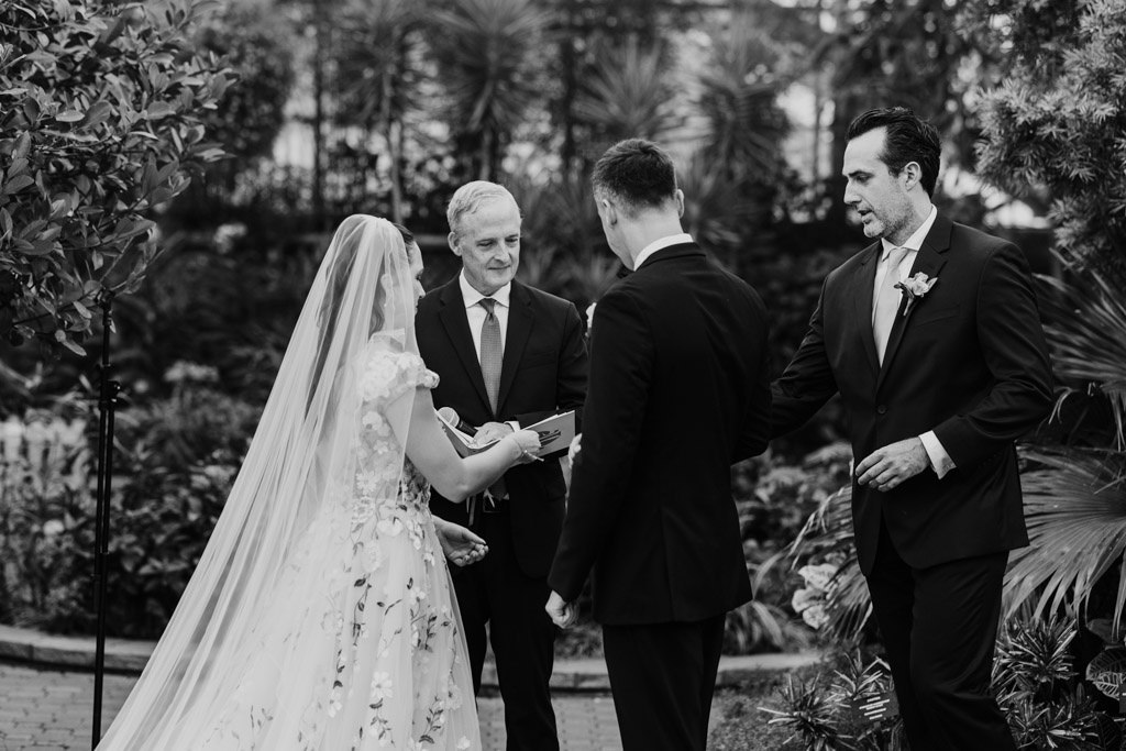 Black and white photo of Best Man handing rings to the groom during Lincoln Park Conservatory wedding ceremony