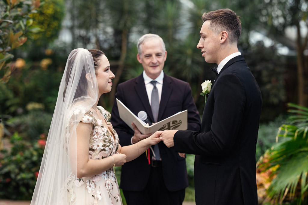 Bride and groom exchange rings during a beautiful Lincoln Park Conservatory wedding ceremony with an officiant standing behind them