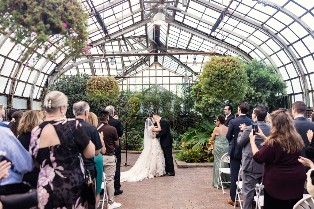 Bride and groom share their first kiss at their Lincoln Park Conservatory wedding ceremony as guests look on and applaud
