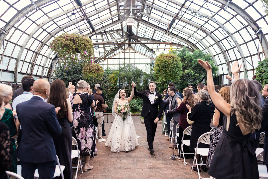 Newlywed couple walks down the aisle in the bright Lincoln Park Conservatory wedding greenhouse as guests cheer and clap around them