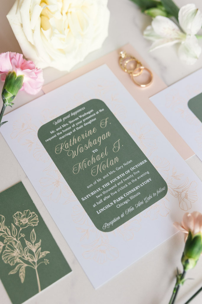 Wedding invitation with gold rings, flowers, and green floral accents on a light surface for Lincoln Park Conservatory wedding ceremony