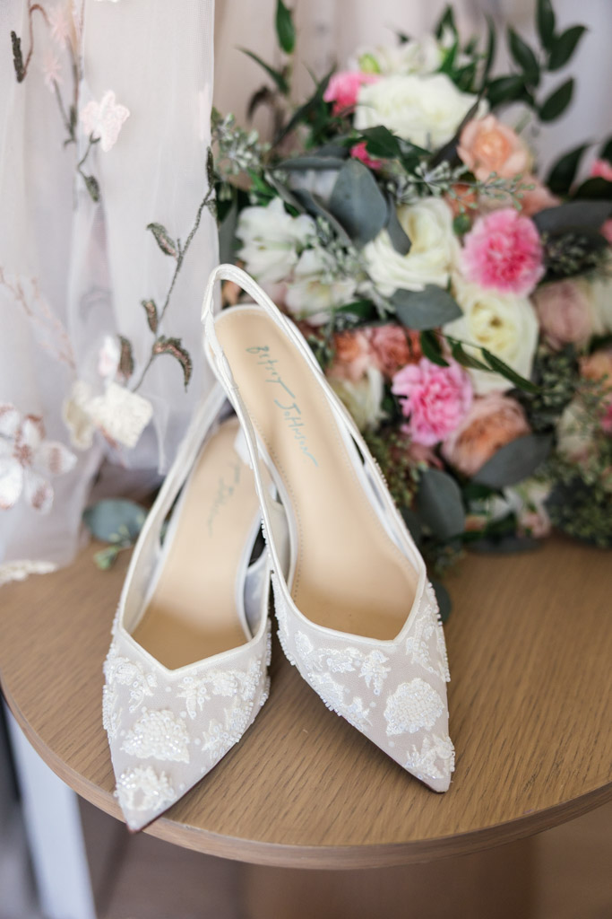 Bride's embroidered white bridal shoes rest on a wooden table, with a bouquet of pink and white flowers in the background