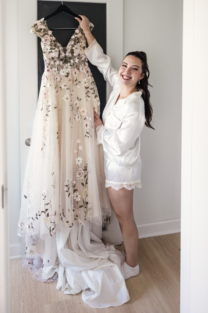 Smiling bride in white robe holds up a floral wedding dress on a black hanger, preparing for her Lincoln Park Conservatory wedding in a bright room