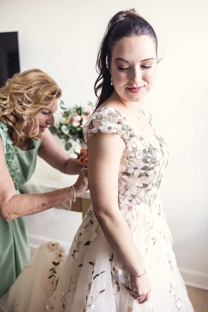 Bride smiles as her mother helps her with the back of her floral wedding dress, preparing for a Lincoln Park Conservatory wedding
