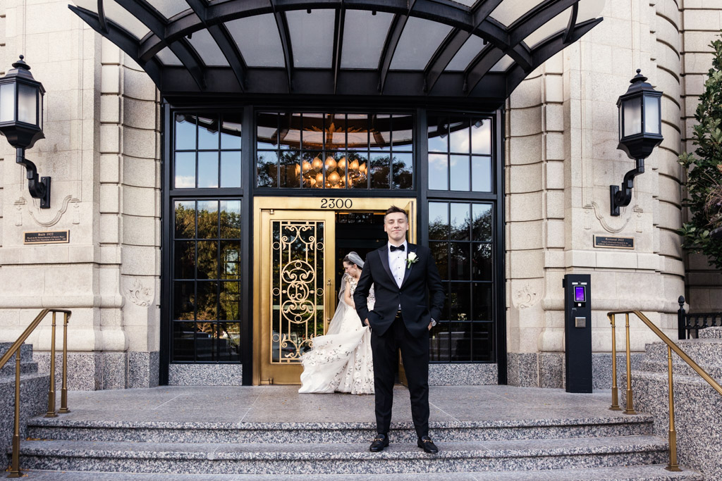 A groom in a tuxedo stands on steps in front of an ornate golden door at The Belden-Stratford, with the bride in a walking up behind him during their first look before their Lincoln Park Conservatory wedding ceremony