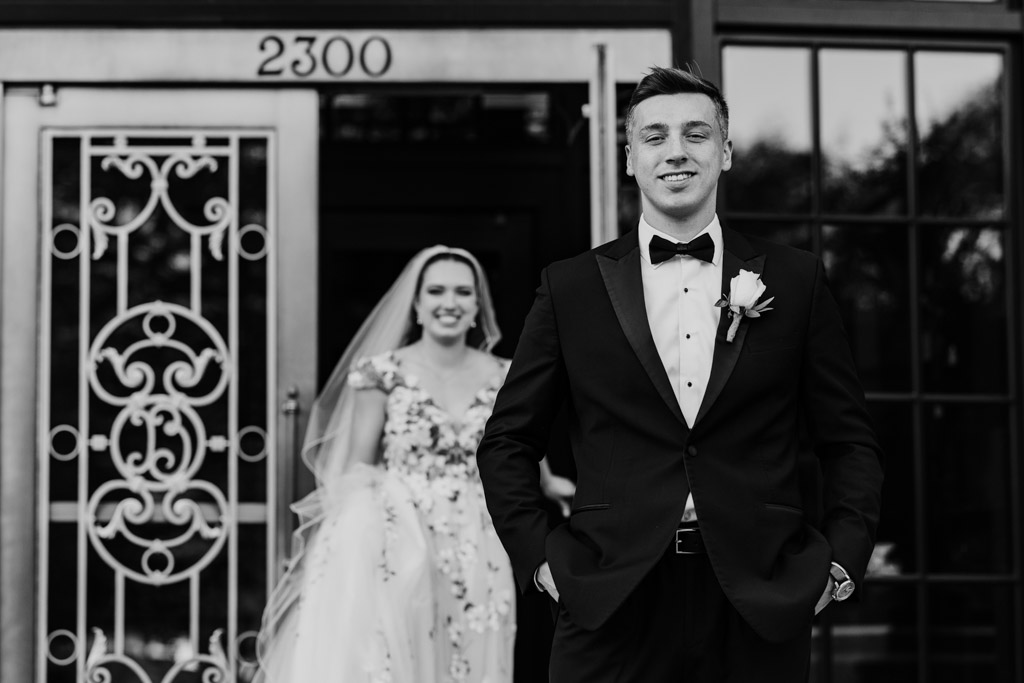 Black and white photo of groom in a tuxedo as the bride approaches during their first look in front of an ornate door before their Lincoln Park Conservatory wedding ceremony