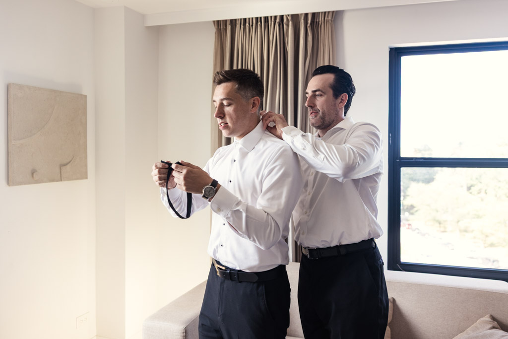 Best Man helps groom with his collar and bow tie as they get ready at Hotel Lincoln before Lincoln Park Conservatory wedding ceremony