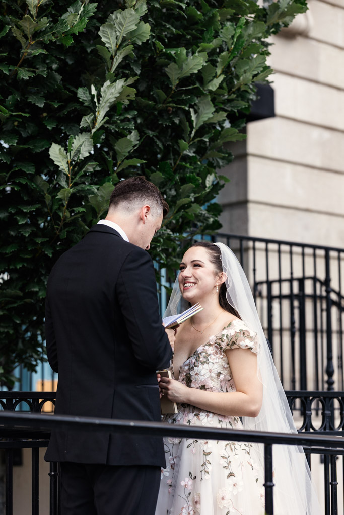 Groom reads vows to bride outdoors by a black railing and green tree outside The Belden-Stratford in Chicago