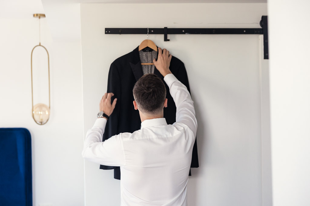 Groom in a white shirt reaching for a black suit jacket hanging on a rack, preparing for his Lincoln Park Conservatory wedding