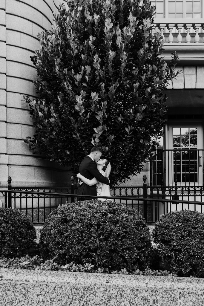 Black and white photo of bride and groom kissing under a large tree in front of The Belden-Stratford before their Lincoln Park Conservatory wedding