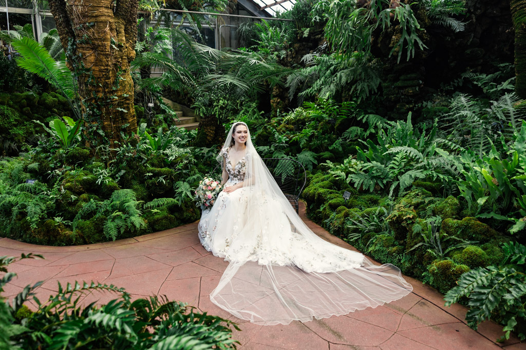 Bride in a white floral dress and long veil sits in a lush, green indoor garden surrounded by ferns and plants at Lincoln Park Conservatory