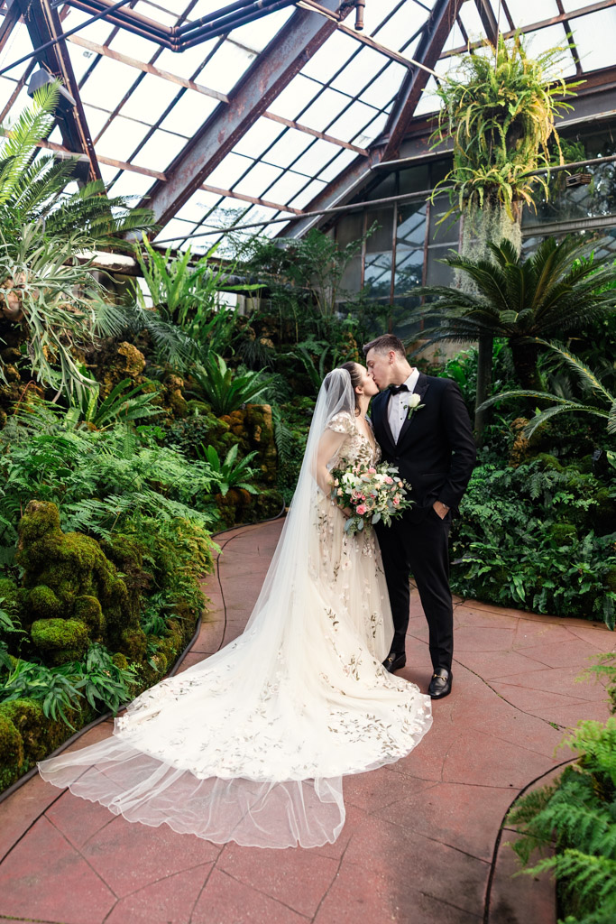 A bride and groom kiss in a lush greenhouse, surrounded by greenery and sunlight before their Lincoln Park Conservatory wedding ceremony