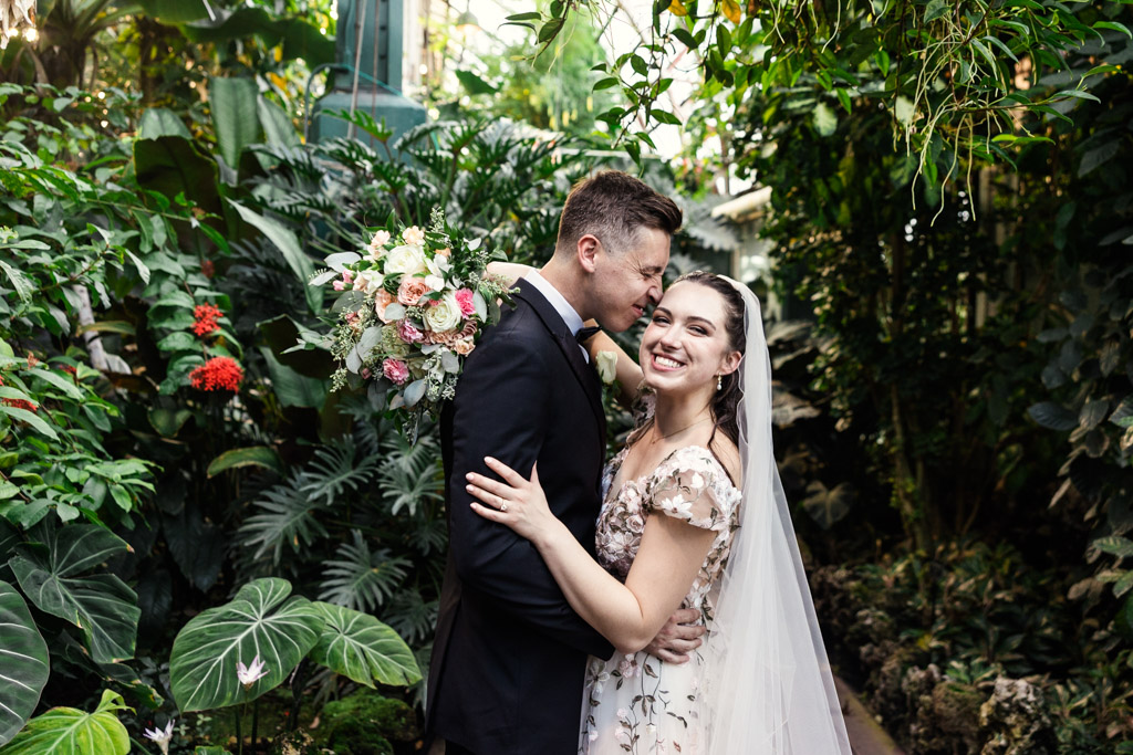 Bride and groom smiling and embracing in a lush, green garden before their Lincoln Park Conservatory wedding ceremony, holding a beautiful bouquet of flowers.