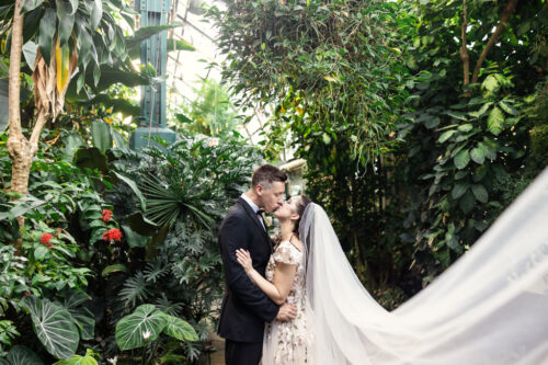 A bride and groom kiss in the lush Lincoln Park Conservatory, surrounded by tropical plants and a flowing bridal veil.