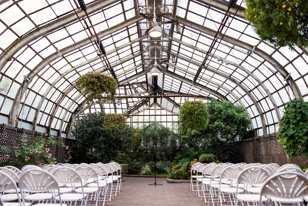 Wedding ceremony space at Lincoln Park Conservatory with rows of white chairs facing a microphone in a glass greenhouse filled with lush greenery