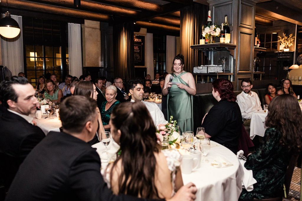 Maid of Honor in a green dress gives a speech to seated guests at wedding reception at Mon Ami Gabi restaurant