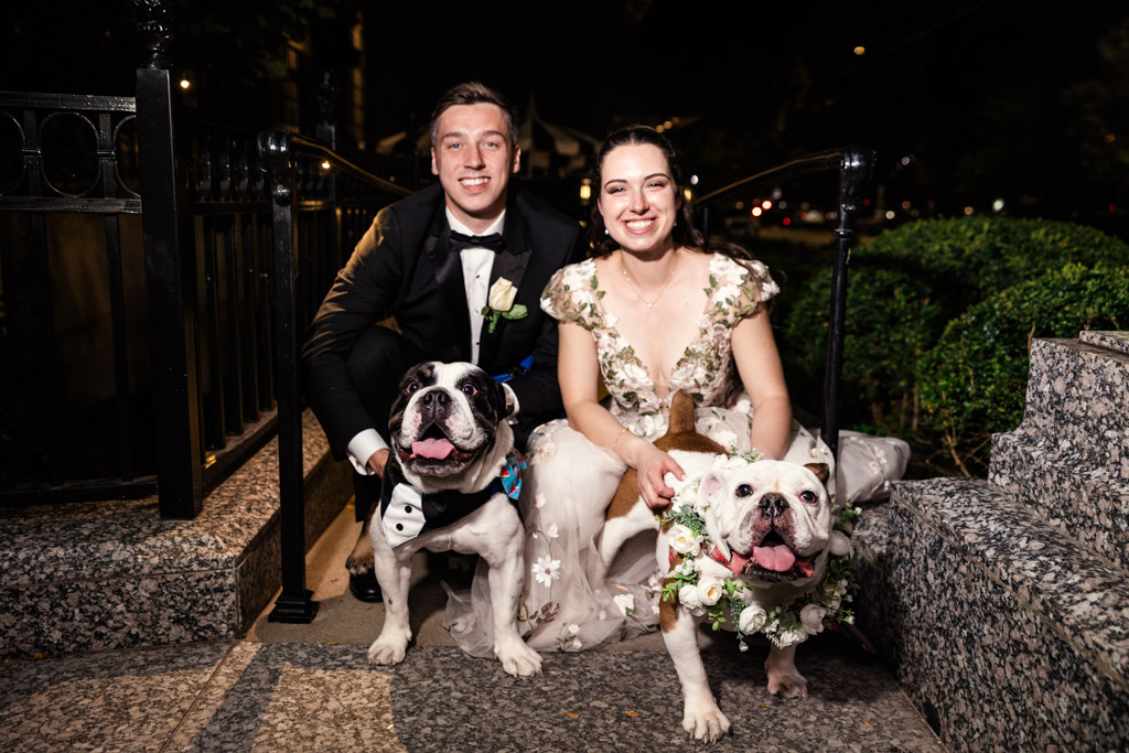 A smiling couple in formal attire pose outdoors at night with two bulldogs wearing floral collars, capturing the charm of a Lincoln Park Conservatory wedding.