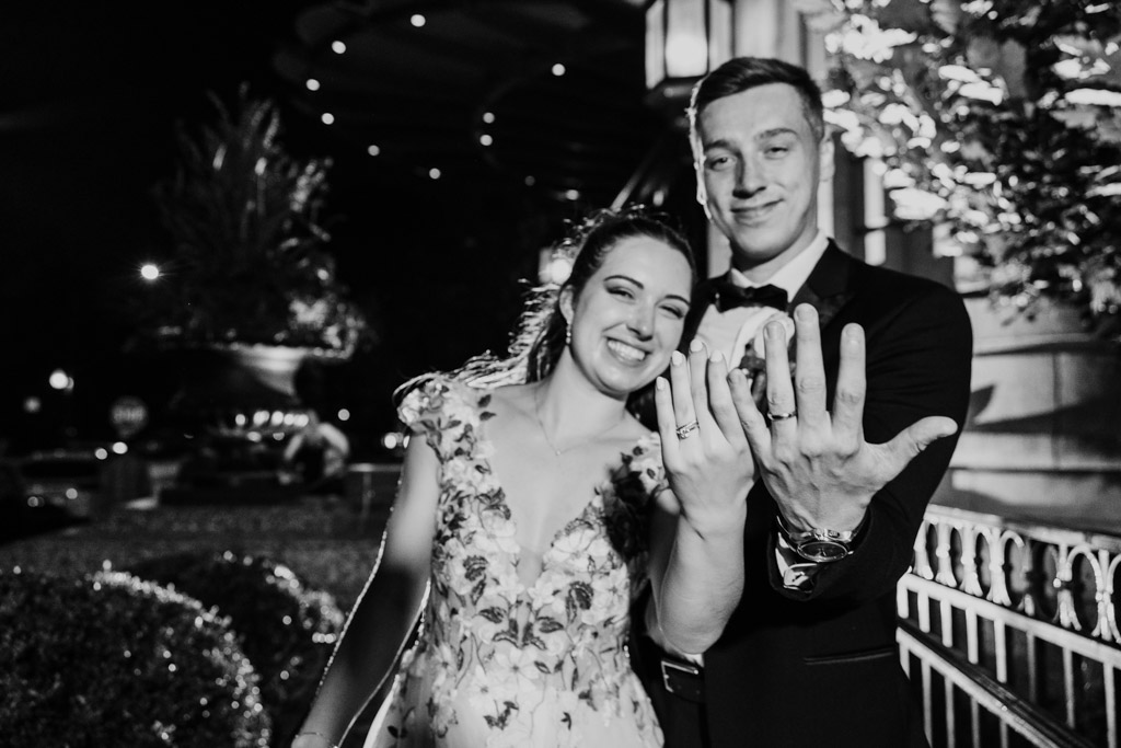 Black and white photo of smiling couple in wedding attire shows their rings to the camera at night outside their Mon Ami Gabi wedding reception