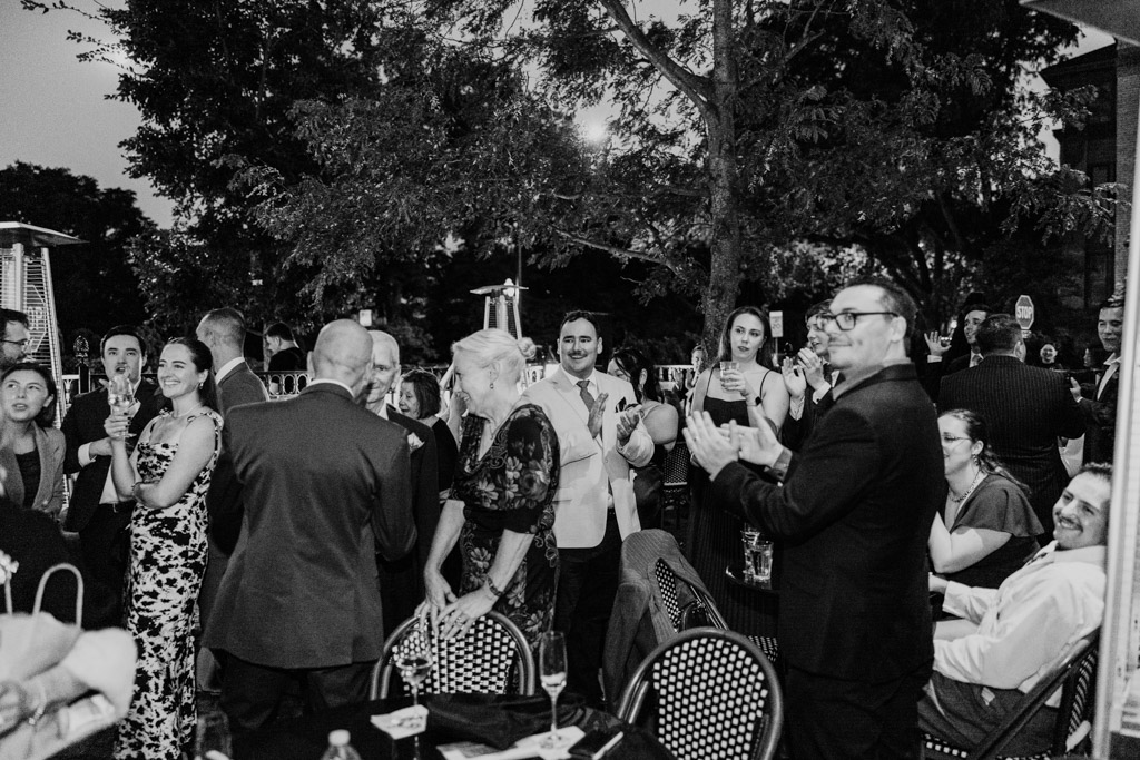 Black and white photo of guests dressed formally gather outdoors at night, smiling and clapping as the newlyweds enter cocktail hour at Mon Ami Gabi in Chicago