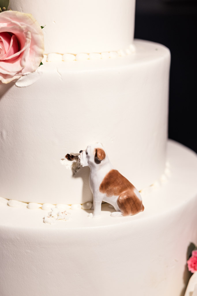 A figurine of a dog appears to bite into a white tiered wedding cake decorated with flowers for wedding reception at Mon Ami Gabi