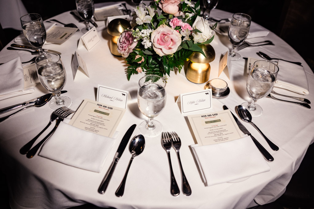 Guest table setting for wedding reception at Mon Ami Gabi featuring menus, place cards, water glasses, and a floral centerpiece atop a crisp white tablecloth