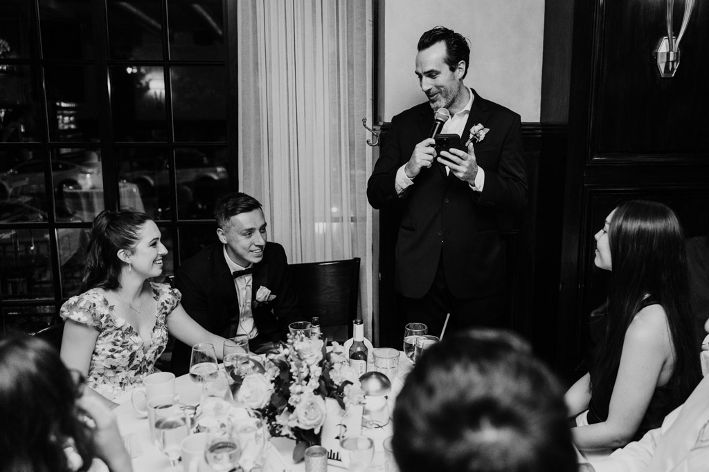 Black and white photo of Best Man giving a speech with a microphone at a table, while the newlyweds and other guests seated smile and listen