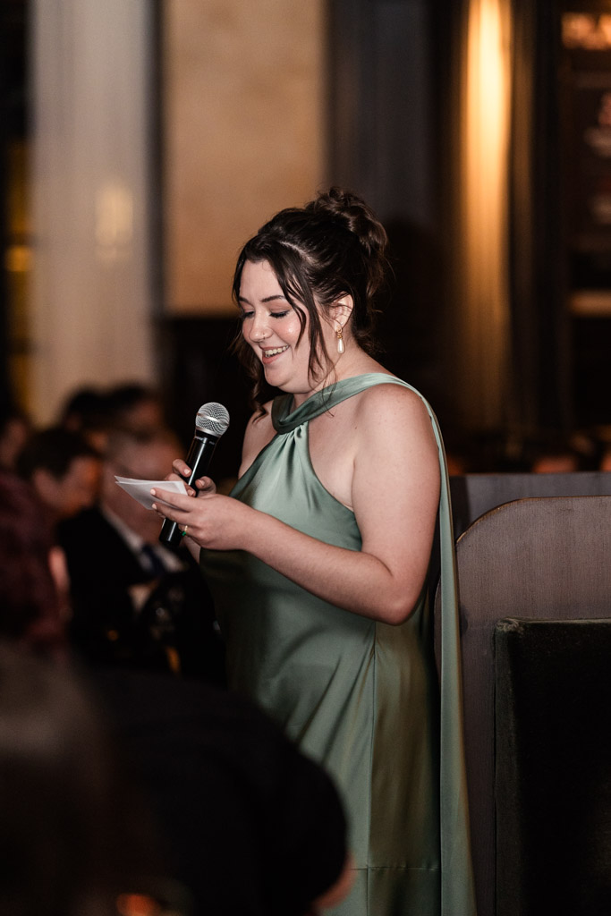 Maid of Honor in a green dress smiles while speaking into a microphone during wedding reception at Mon Ami Gabi