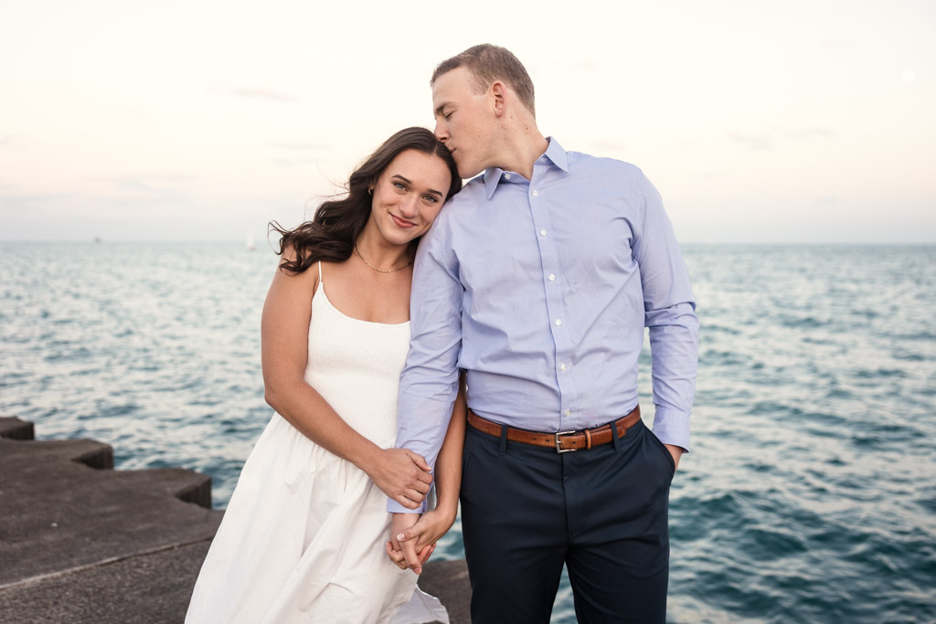 Man kisses his partner's head as they stand holding hands by Lake Michigan during their Montrose Harbor engagement session