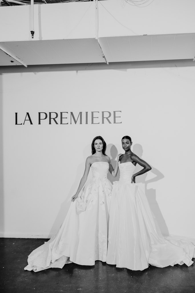 Two women in elegant white gowns stand in front of a wall with the words 