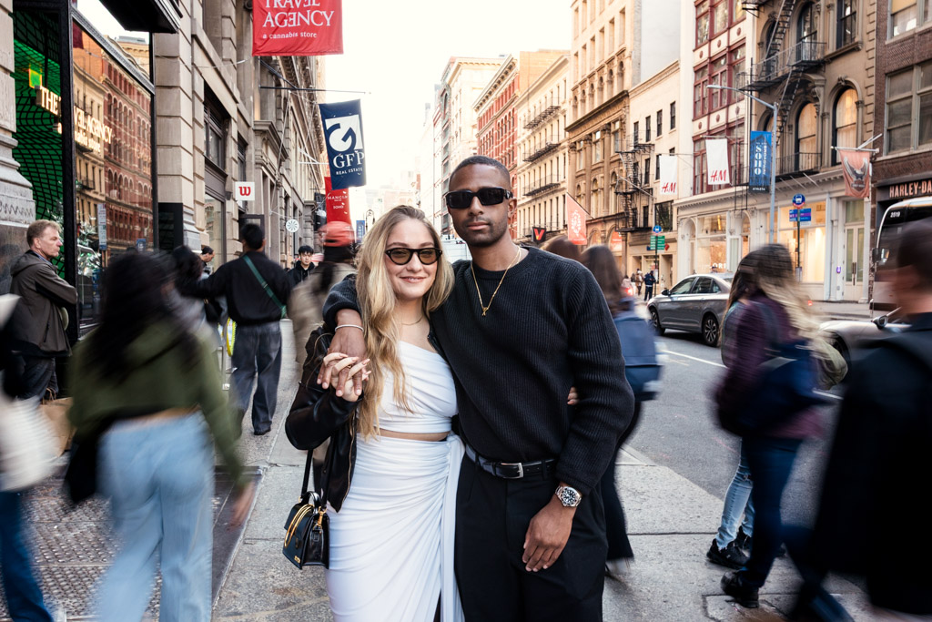 A stylish couple poses together on a busy city street, with people walking by in the background.